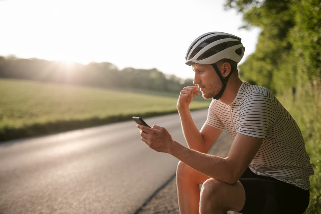ciclista_pensativo_sentado_con_bicicleta_en_camino_rural_al_atardecer_revisando_móvil